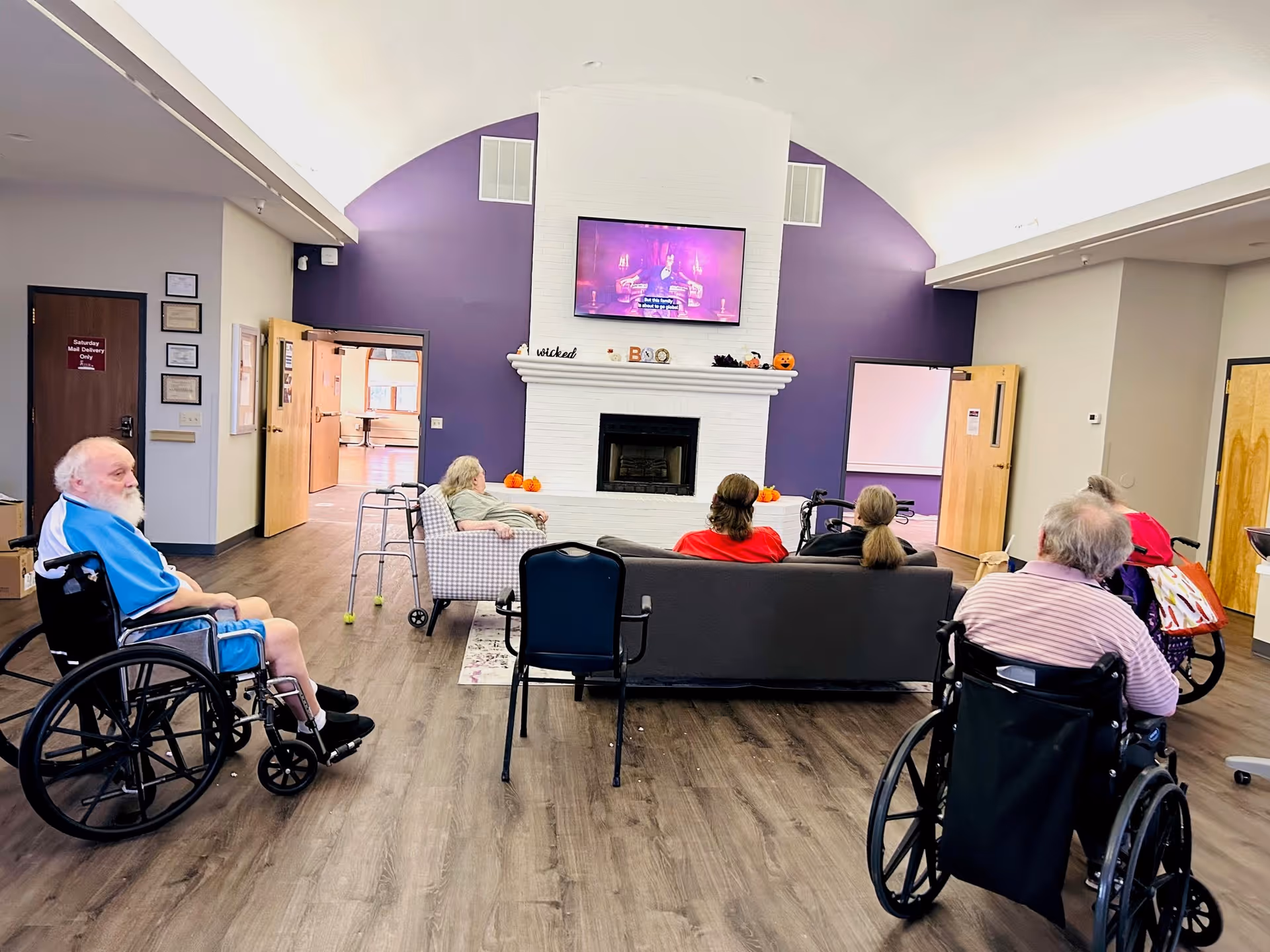 A group of elderly individuals, some in wheelchairs, sitting in a common area with a high vaulted ceiling and purple accent walls, watching a television mounted above a white brick fireplace decorated with small pumpkins and Halloween-themed items.