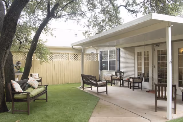 Outdoor patio area at Brookdale North Austin with wooden benches and chairs arranged on a concrete and artificial grass surface, shaded by a large tree and a covered porch attached to the building.