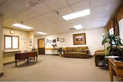 Spacious senior care facility lobby with sofas, a small desk, framed artwork, plants, and fluorescent ceiling lights.
