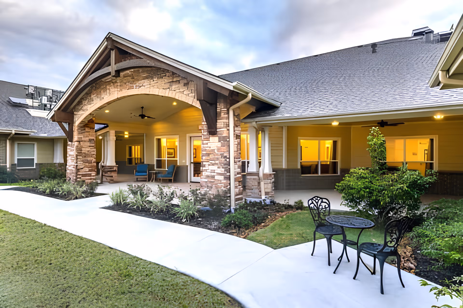 Outdoor patio area of Discovery Village Clear Lake featuring a covered seating area with two blue chairs and a small table under a stone archway. There is a paved walkway, green lawn, and a small black metal table with two chairs near some bushes. The building exterior is light-colored with large windows and warm indoor lighting visible.