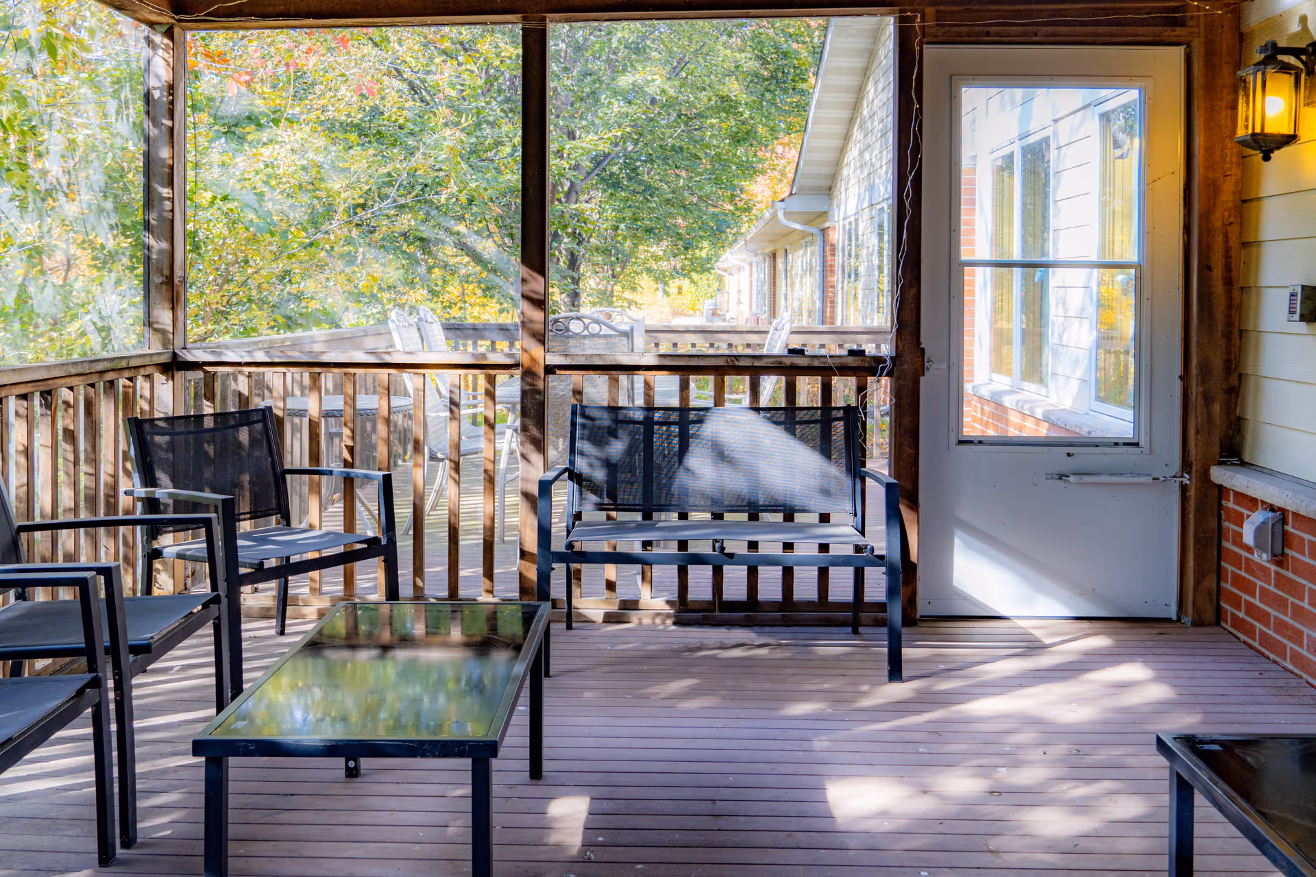 A screened-in porch area with black metal chairs and a matching bench around a glass-top coffee table. The porch has wooden flooring and railing, with a white door leading inside. Outside the porch, there are trees with green leaves and a row of similar building units visible.