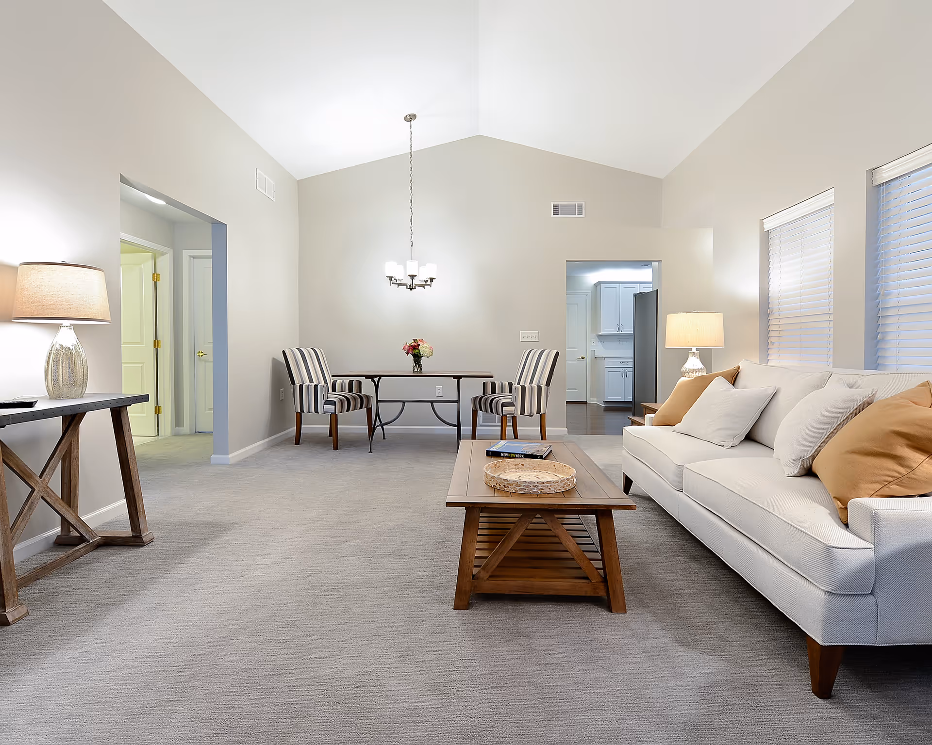 Bright, open living room with a white sofa, wooden coffee table, striped dining chairs at a small table, and neutral walls and carpeting.