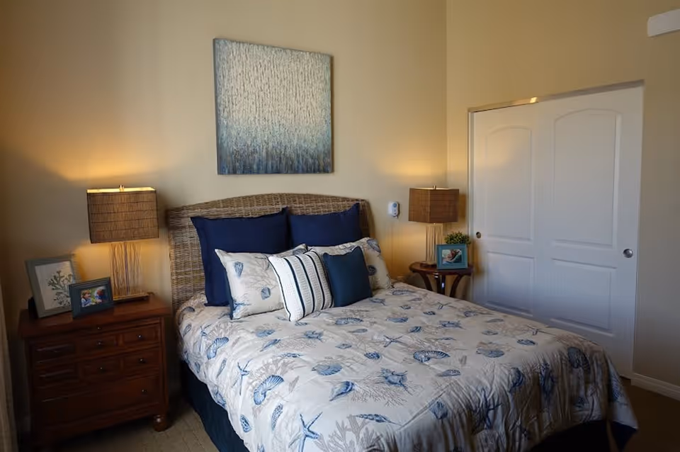 A cozy bedroom with a bed featuring a seashell-patterned comforter and multiple pillows in shades of blue and white. The bed has a wicker headboard. On either side of the bed are wooden nightstands with matching lamps that have rectangular shades. There are framed pictures and a small plant on the nightstands. A piece of abstract art hangs on the wall above the bed, and there is a white double-door closet on the right side of the room.