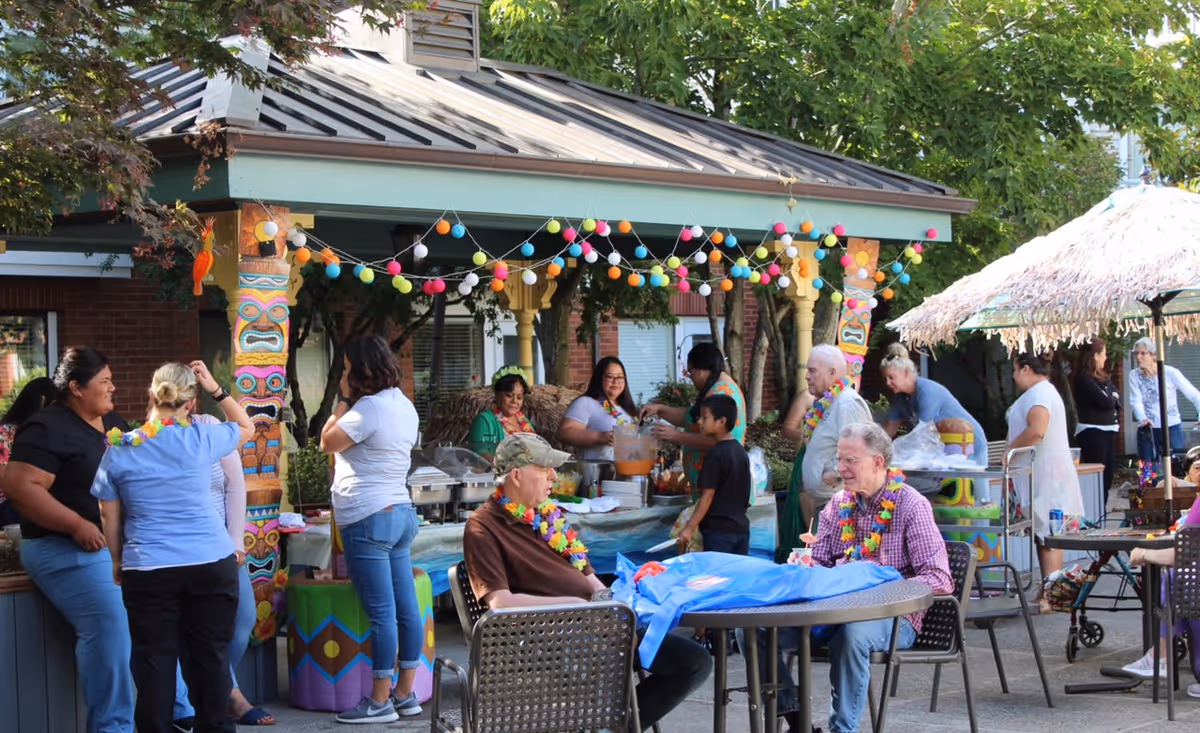 People socializing at a decorated outdoor pavilion with a buffet, tiki-style decorations, and tables with seated guests.