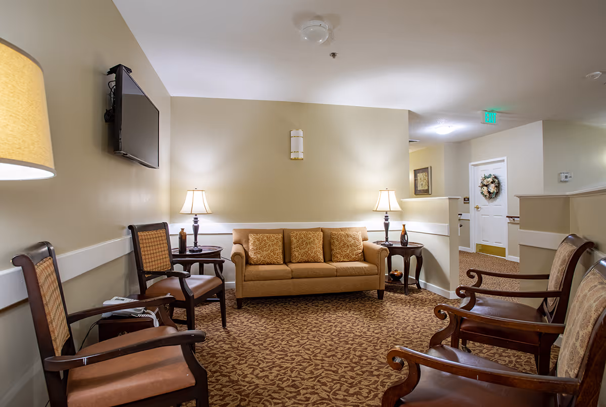A cozy common sitting area with a sofa, several wooden armchairs, side tables, lamps and a wall-mounted TV in a senior living facility.