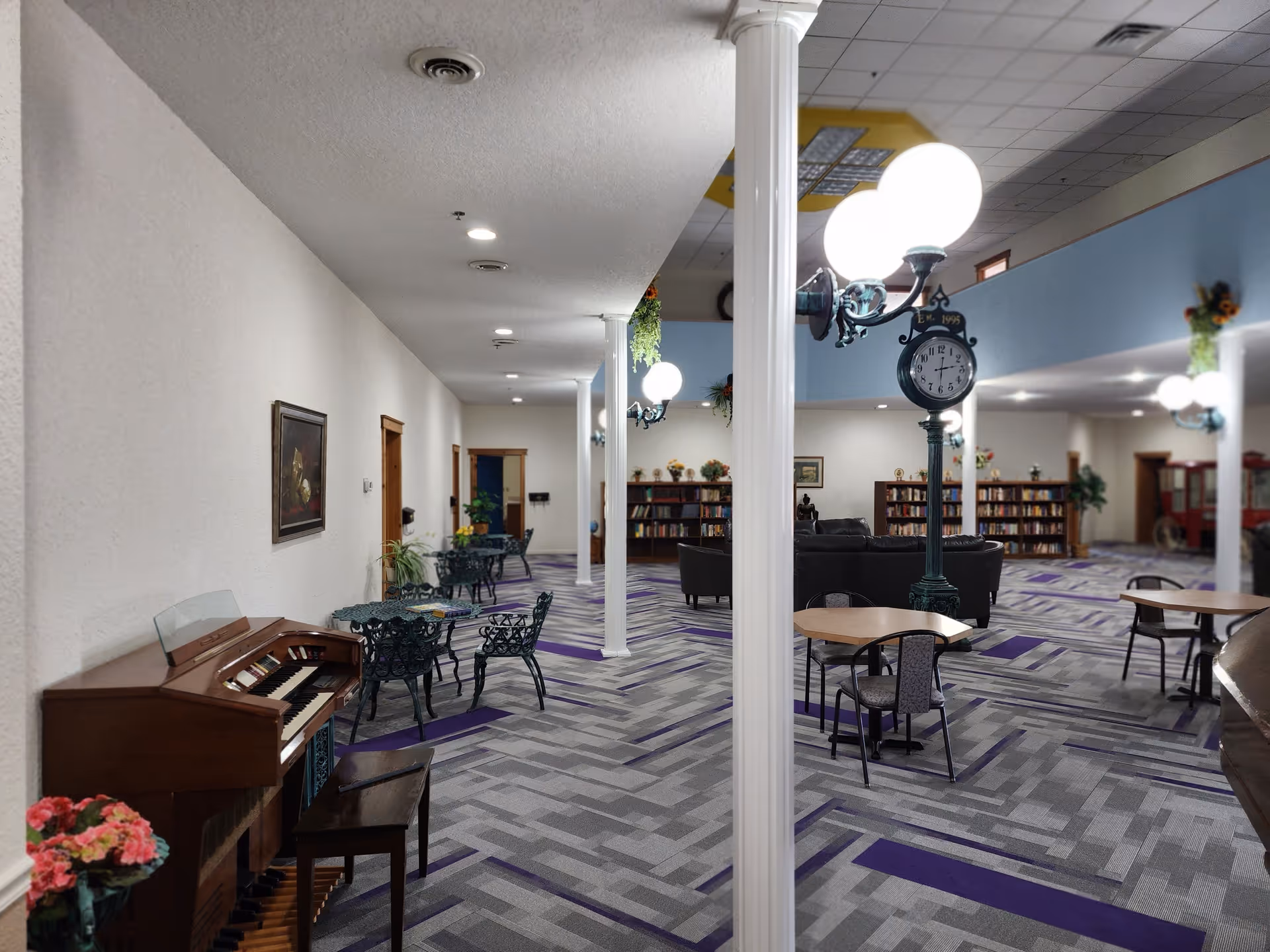 A spacious common area in an assisted living facility featuring a patterned carpet with purple accents, white columns, round tables with chairs, a vintage-style standing clock, bookshelves filled with books, a piano with a bench, and decorative plants and paintings on the walls.