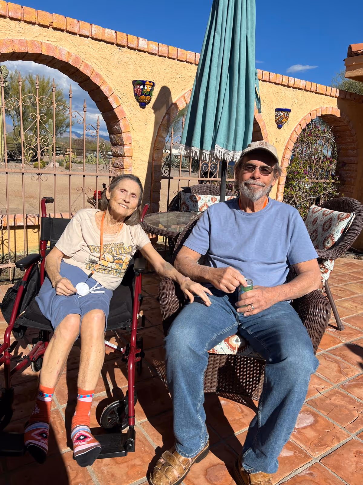 An elderly woman sitting in a wheelchair and an elderly man sitting in a wicker chair on a sunny outdoor patio with terracotta tiles and a yellow stucco wall with brick arches in the background. The woman is smiling and reaching out to the man, who is wearing sunglasses and a cap, holding a can. There is a green patio umbrella and decorative wall sconces on the wall behind them.