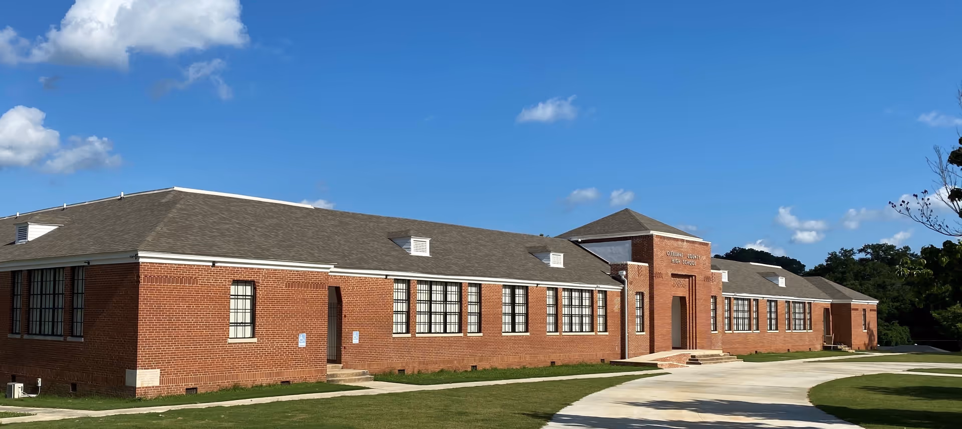 Exterior view of a large single-story brick building with multiple windows and a central entrance under a clear blue sky with scattered clouds. The building is surrounded by a well-maintained lawn and a curved concrete driveway.