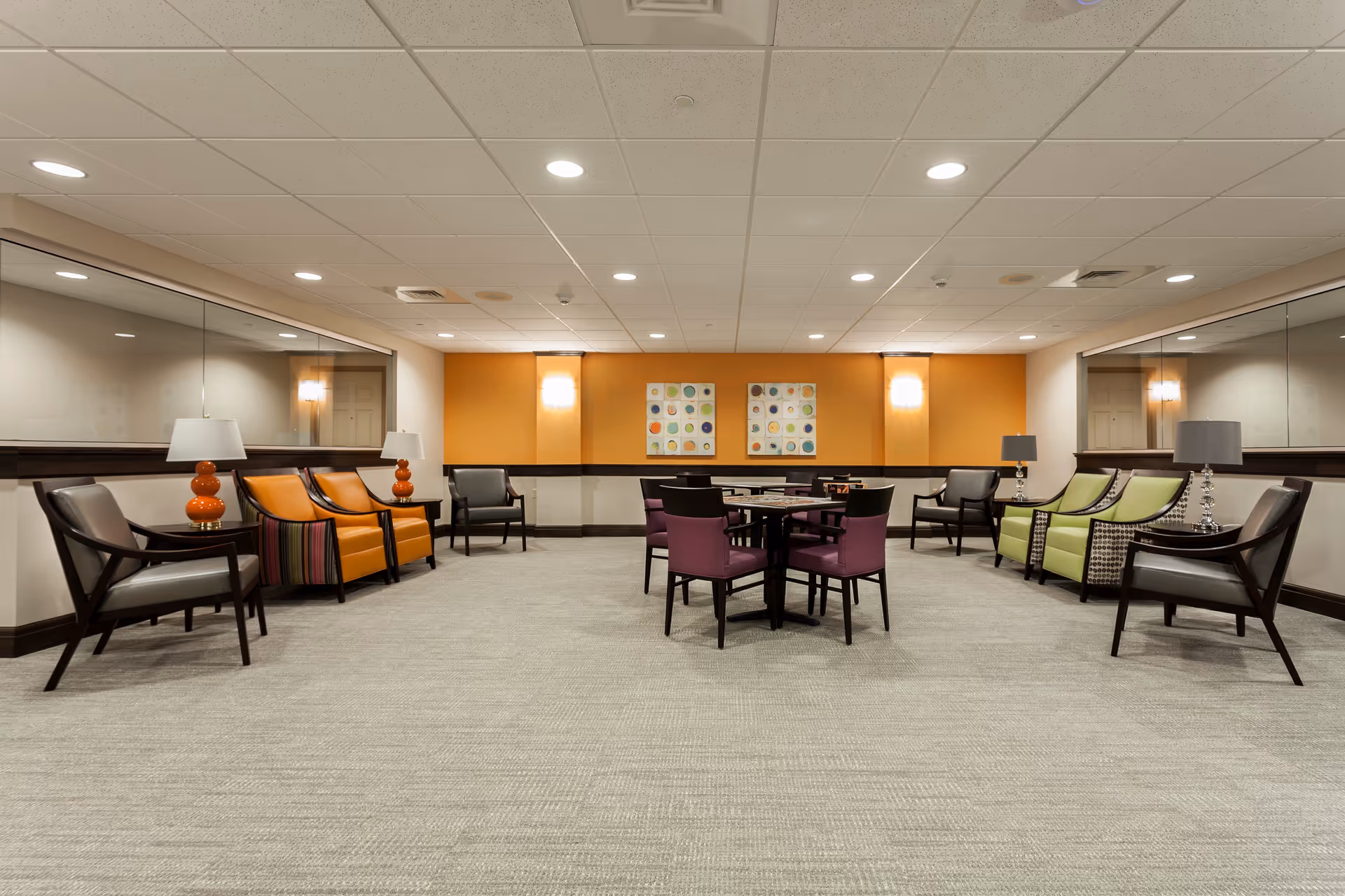 A spacious, well-lit common area with a neutral carpeted floor and a white ceiling with recessed lighting. The room features a variety of chairs in different colors including orange, green, gray, and purple, arranged around small tables. There are two colorful abstract paintings on an orange accent wall at the far end. Large mirrors line the side walls, and table lamps with orange and gray bases are placed on side tables next to some chairs.