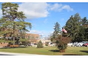Outdoor view of a senior living facility with a lawn, trees, parked cars, and an American flag on a flagpole under a partly cloudy sky.