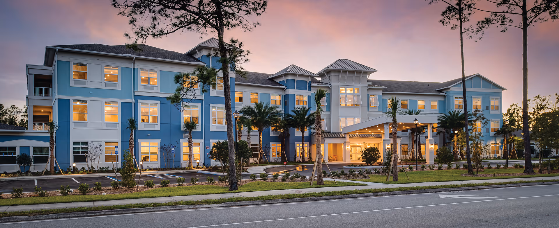 Exterior view of a large, three-story senior living facility building named Sancerre at Palm Coast during sunset, with lights glowing from the windows, palm trees and landscaping in front, and a covered entrance area.