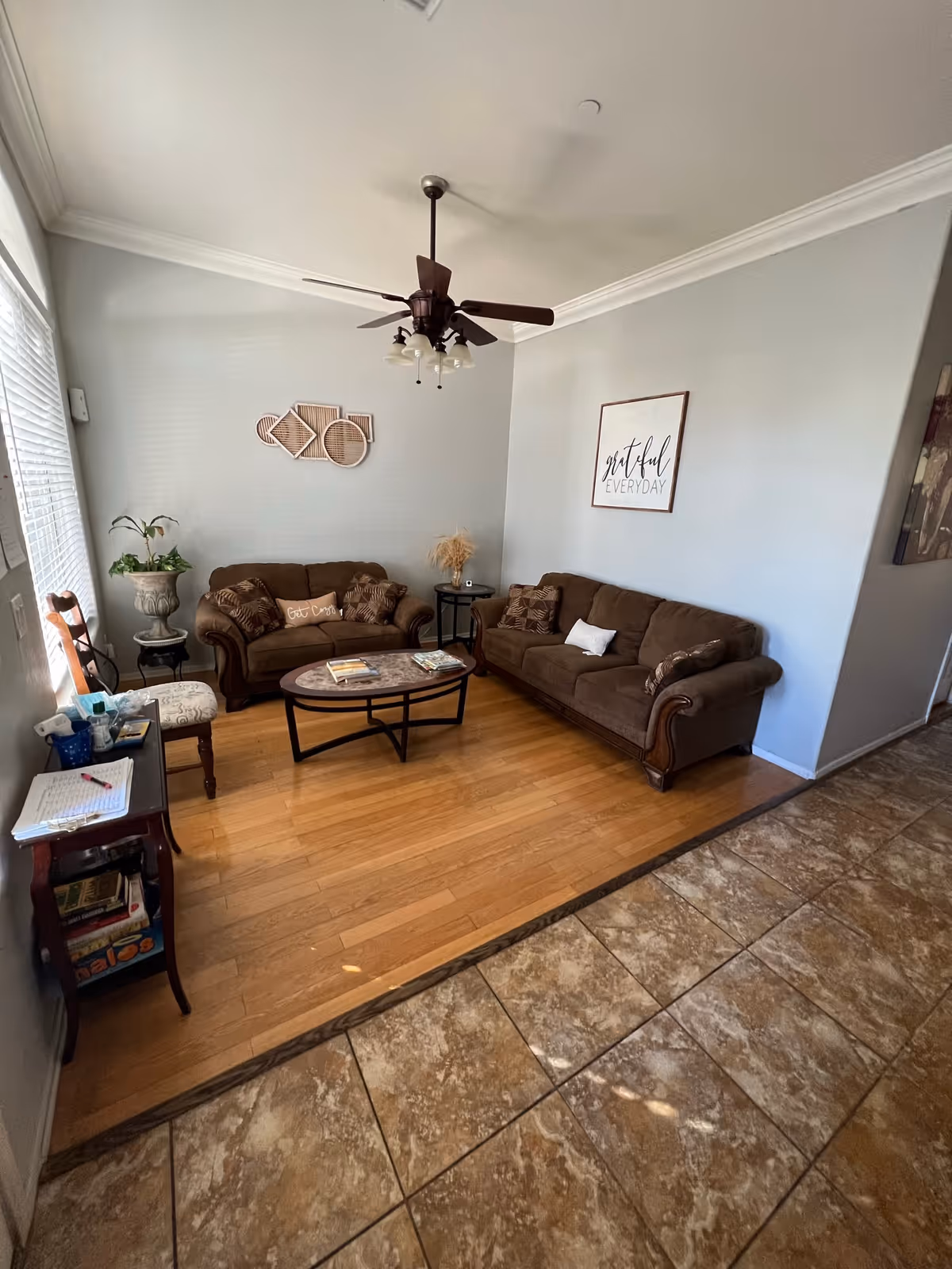 A cozy living room with two brown sofas arranged around an oval coffee table. The room has light blue walls with decorative wall art, including a framed sign that says 'grateful EVERYDAY'. There is a ceiling fan with lights, a small side table with a plant, and a wooden floor area transitioning to a tiled floor. A small table with books and a chair is positioned near a window with blinds.