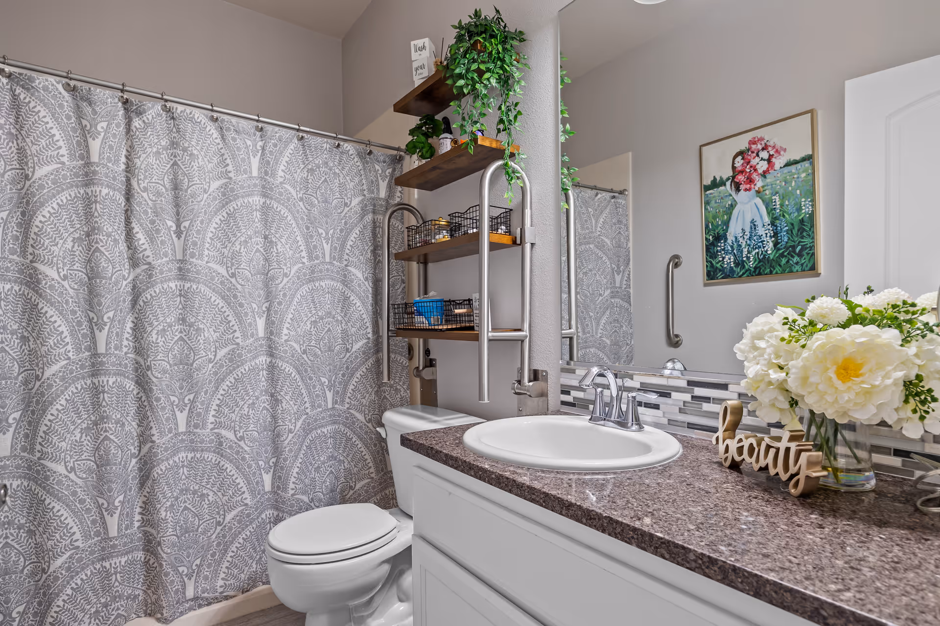 Bathroom with a sink set in a dark granite countertop, toilet, patterned shower curtain, wall shelves, mirror, and floral decor.