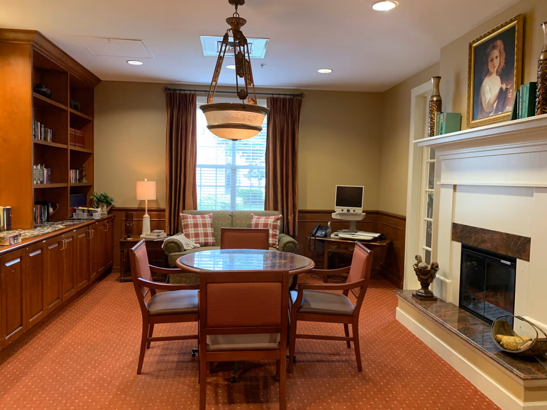 A cozy interior room with a round wooden table surrounded by four chairs in the center. Behind the table is a green couch with two plaid pillows, positioned in front of a window with brown curtains. To the left, there is a built-in wooden bookshelf and cabinet with books and decorative items. On the right side, there is a white fireplace with a marble hearth, decorated with vases, books, and a framed portrait of a young girl. A vintage-style hanging light fixture is above the table, and a small desk with a computer monitor and phone is in the corner.