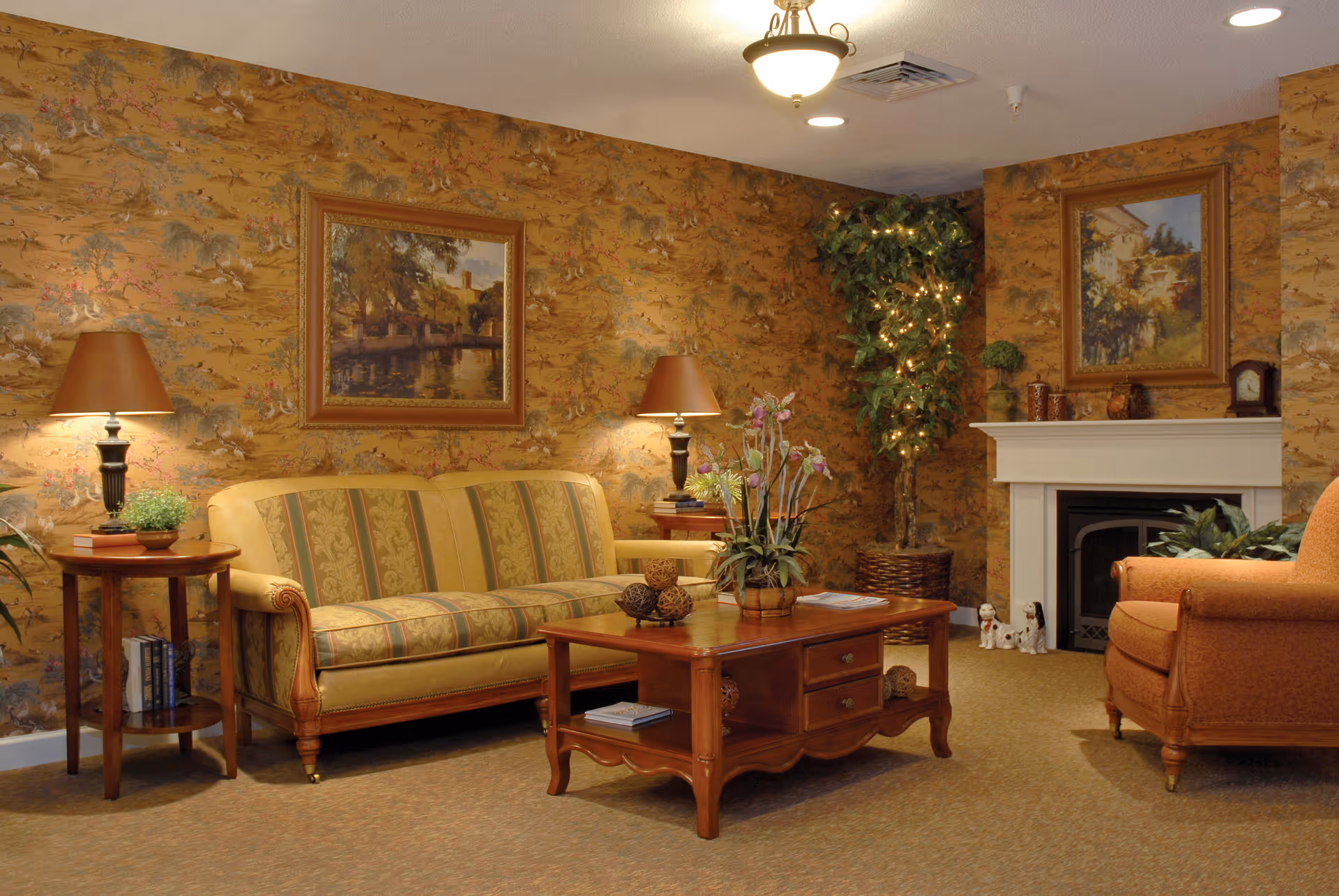 A cozy living room with a patterned yellow and green sofa, a wooden coffee table with decorative items, an orange armchair, two side tables with lamps, a fireplace with framed artwork above it, and a tall potted plant with string lights in the corner. The walls have a warm, patterned wallpaper.
