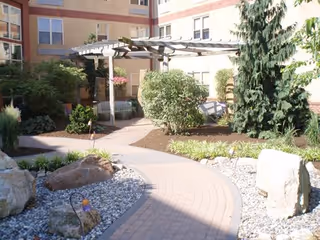 Outdoor courtyard area with a winding brick pathway, surrounded by rocks, bushes, and trees. There is a wooden pergola with benches underneath, adjacent to a multi-story building with windows.