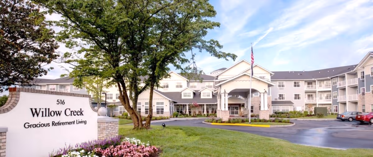 Front exterior of the Willow Creek Gracious Retirement Living building with driveway, entrance canopy, landscaped lawn, and a sign.