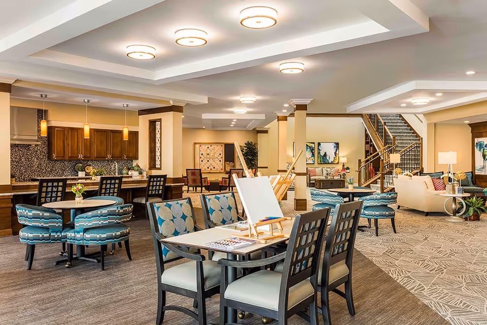 A spacious and well-lit common area in an assisted living facility featuring multiple seating arrangements including round tables with patterned chairs, a table with easels and painting supplies, a kitchen area with wooden cabinets and pendant lights, and a cozy living room area with sofas and artwork on the walls. A staircase is visible in the background.