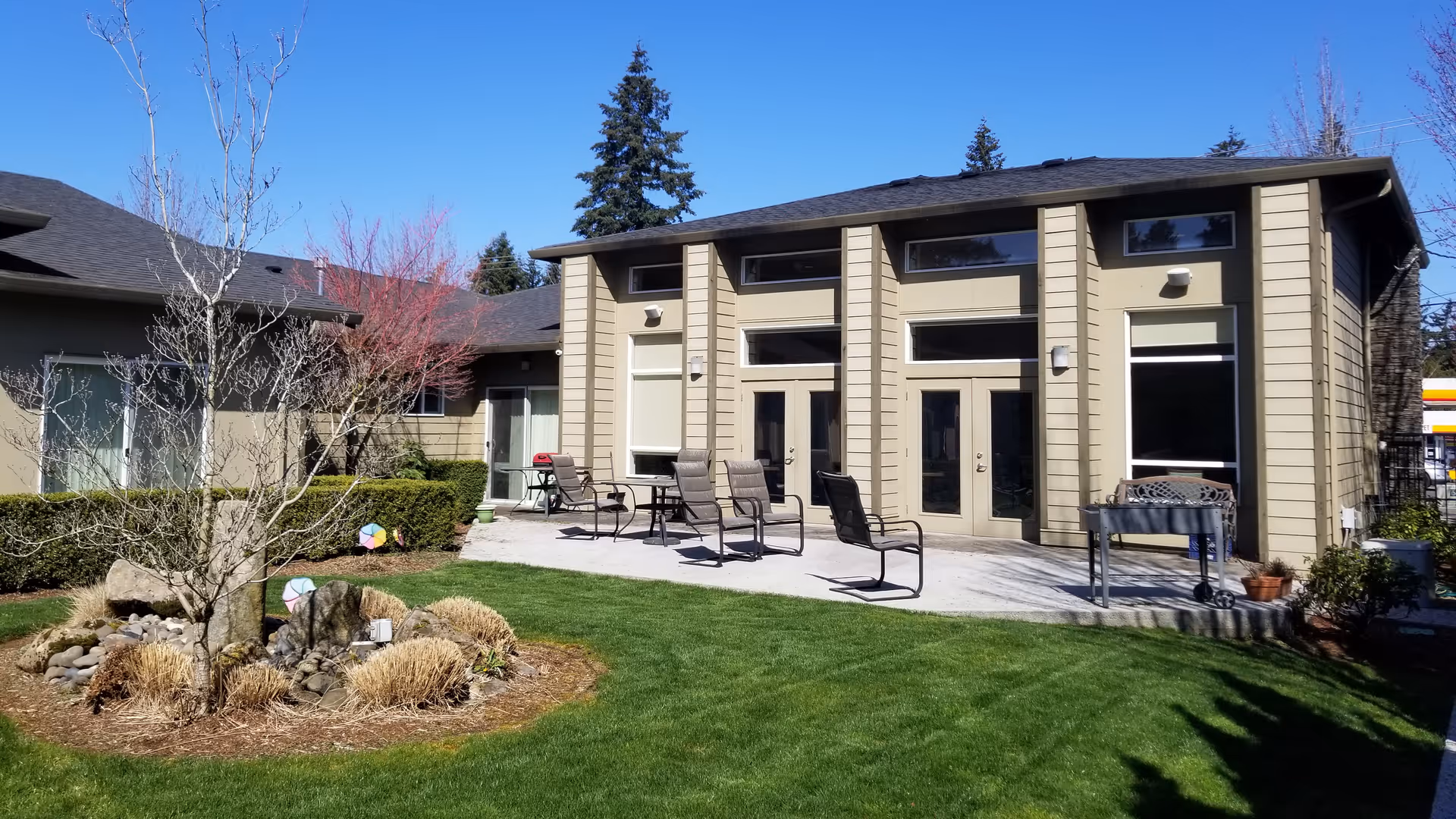 Exterior view of a low, modern beige building with a concrete patio, outdoor chairs, and a manicured lawn and garden.