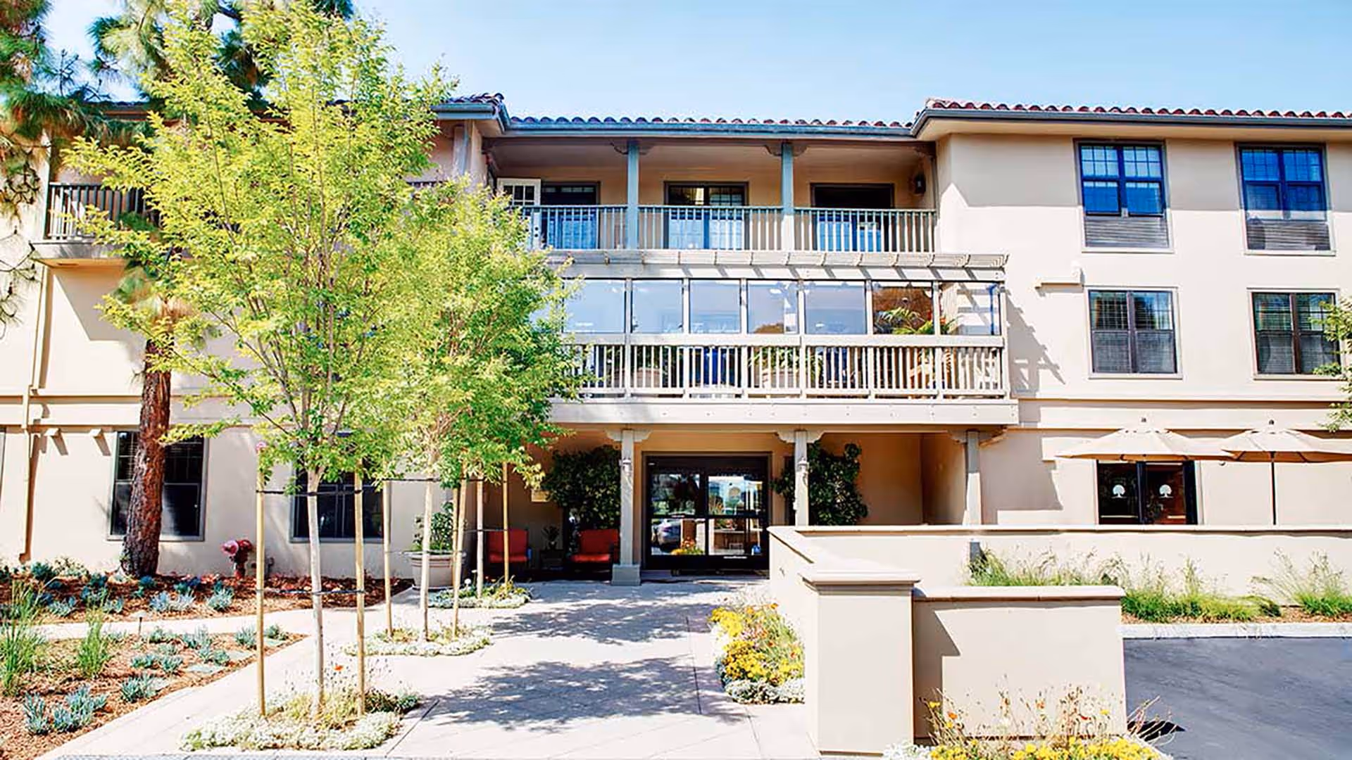 Exterior view of a two-story senior living facility building with beige walls and a tiled roof. The entrance is centered with a covered walkway and glass doors. There are small trees and landscaped plants along the pathway leading to the entrance. Outdoor seating with umbrellas is visible on the right side.