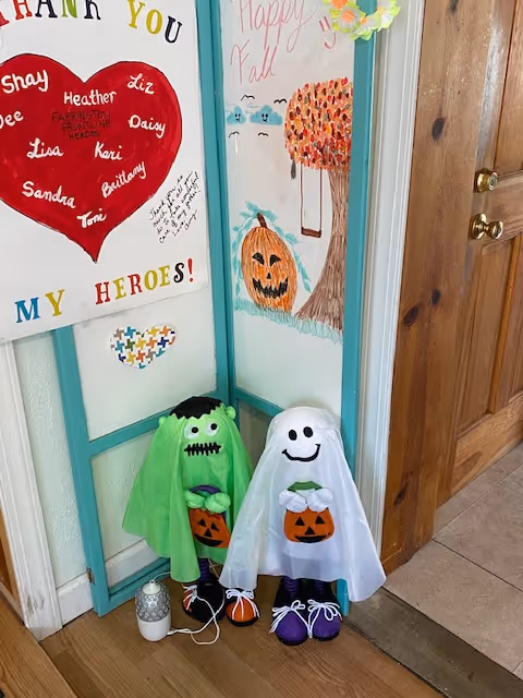 Two Halloween-themed decorations resembling small figures dressed as a green Frankenstein monster and a white ghost, each holding a pumpkin bucket, placed on the floor next to a wooden door. Behind them is a decorative folding screen with colorful drawings including a pumpkin, a tree with autumn leaves, and a red heart with names written inside it. The screen also has the words 'Thank You My Heroes!' and 'Happy Fall' written on it.