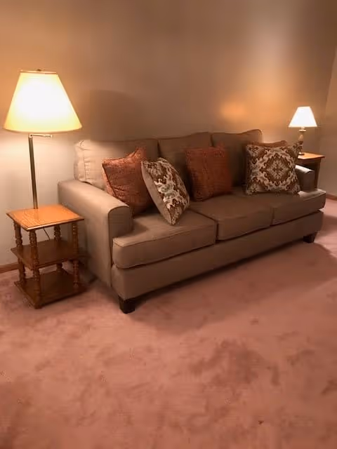 A beige sofa with four decorative pillows in shades of brown and patterned designs is placed against a plain wall. On each side of the sofa, there is a wooden side table with a lamp on top, both lamps are turned on, casting a warm light. The floor is carpeted in a light brown color.