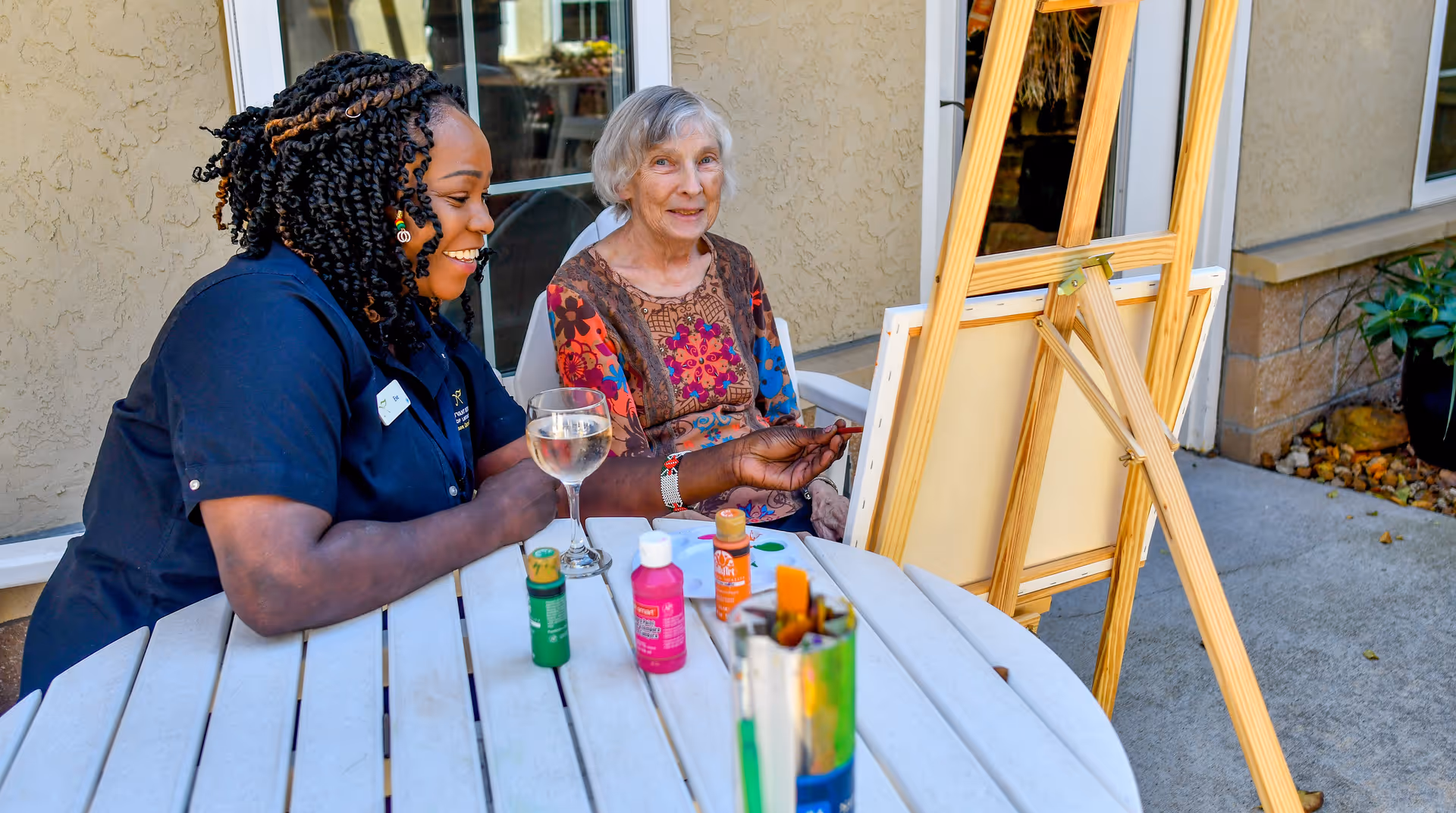 An elderly woman and a caregiver sitting at a round outdoor table with paint bottles and brushes, engaging in a painting activity with blank canvases on easels in front of them. The setting is outside a building with beige walls and windows.