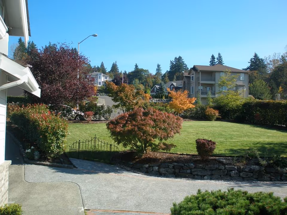 A well-maintained outdoor garden area with green grass, various trees with autumn-colored leaves, shrubs, and a small black metal gate. Residential buildings and a clear blue sky are visible in the background.
