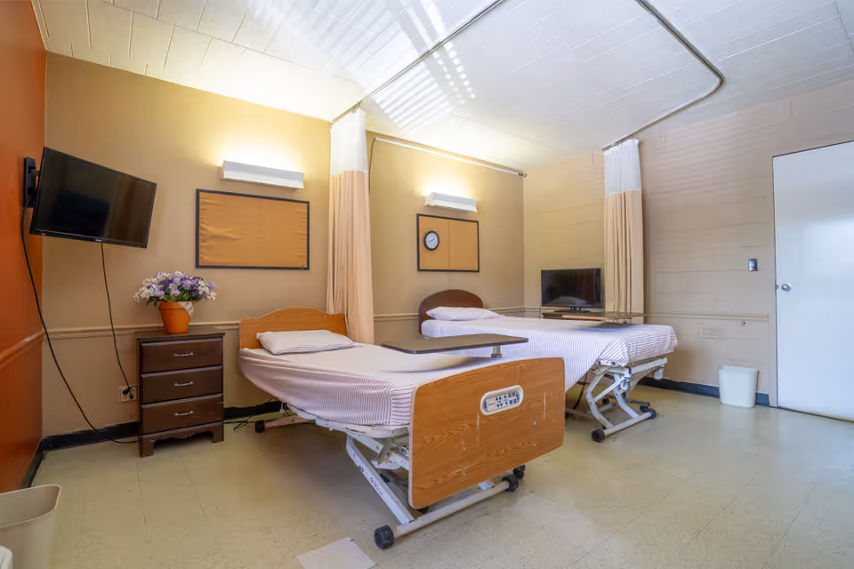 A nursing facility room with two hospital-style beds, privacy curtains, wall-mounted TVs, a bedside dresser with flowers, and a clock on the wall.