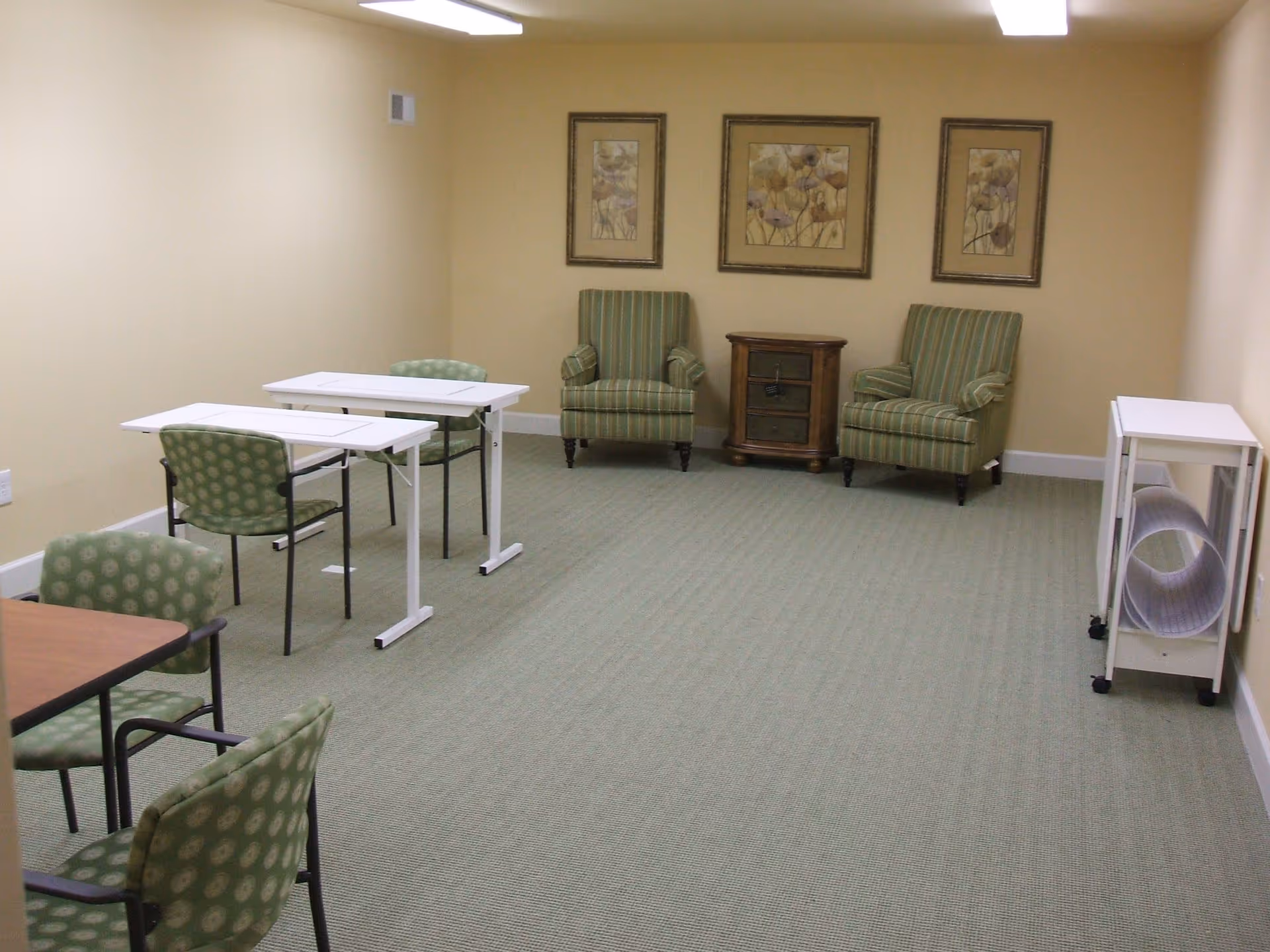 A room with beige walls and green carpet featuring two green striped armchairs with a small wooden side table between them, three framed floral artworks on the wall, two white tables with green patterned chairs, and a white rolling cart with a rolled-up mat on the right side.