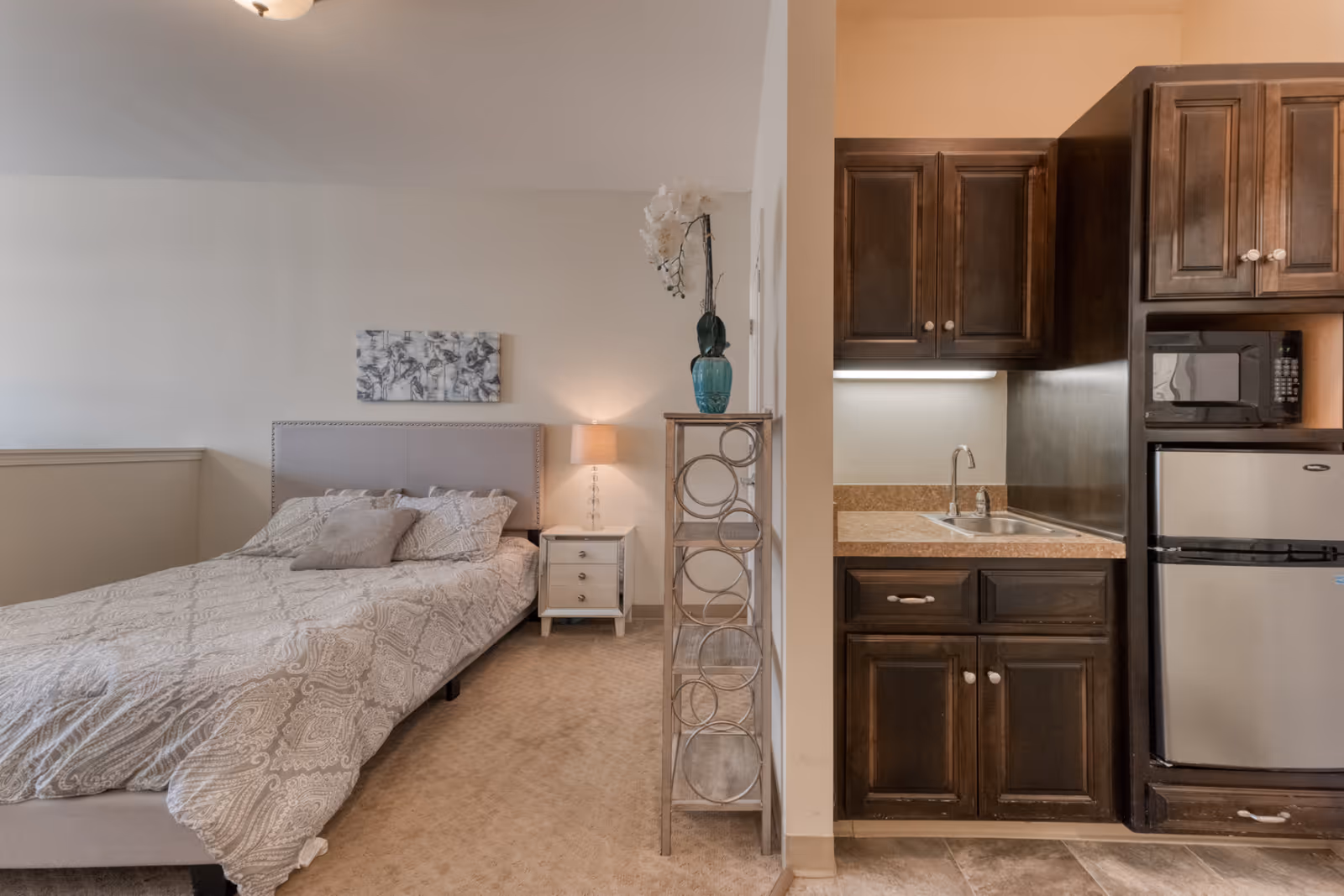 A cozy studio apartment featuring a bed with patterned bedding and pillows on the left side, a nightstand with a lamp, and a decorative painting on the wall above the bed. On the right side, there is a small kitchenette with dark wooden cabinets, a sink, a microwave, and a mini refrigerator. A decorative shelving unit with a vase and flowers separates the sleeping area from the kitchenette.