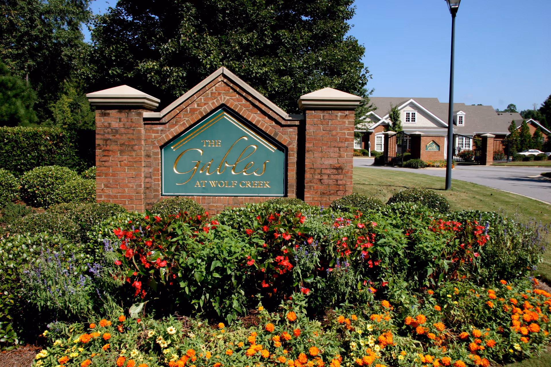 Brick entrance sign for The Gables at Wolf Creek surrounded by colorful flowers and greenery, with a paved road and residential buildings in the background under a clear blue sky.