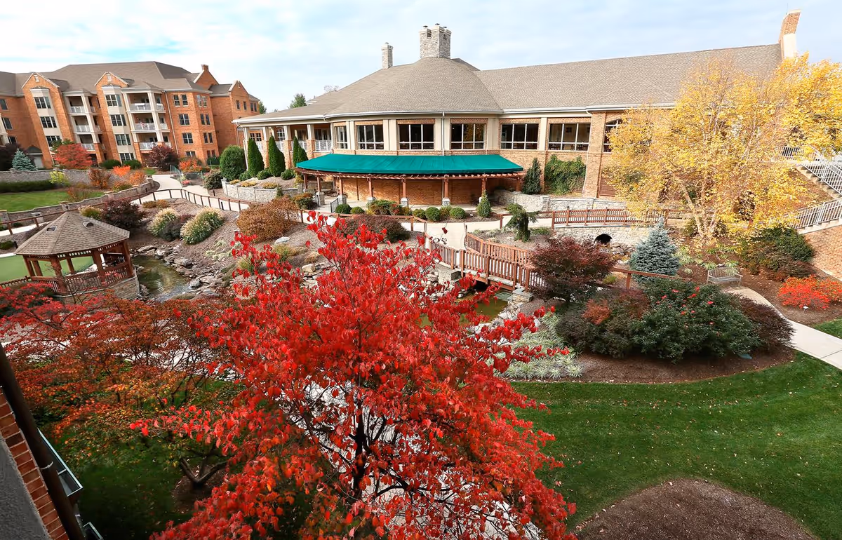View of an outdoor garden area at Shenandoah Valley Westminster-Canterbury Life Plan Community featuring a variety of trees with autumn foliage, a small wooden gazebo over a pond, a wooden bridge, and a large brick building with a green awning in the background.