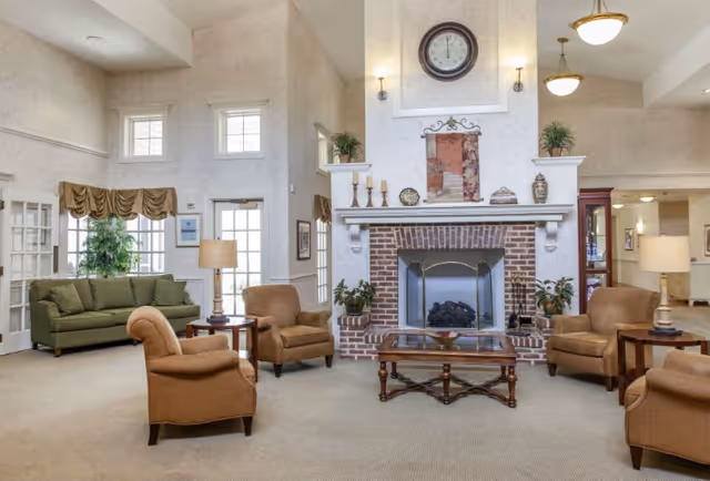 A spacious senior living facility common area with a large brick fireplace as the focal point. The room features beige walls and carpet, a green sofa near windows with valances, four brown armchairs arranged around a wooden coffee table, and two side tables with lamps. Above the fireplace is a large clock and decorative items. The room is well-lit with natural light from windows and ceiling lights.