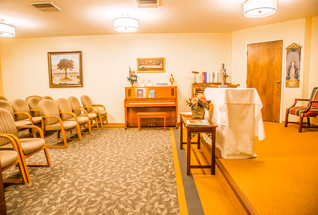 A small chapel or prayer room with rows of beige chairs on the left, a wooden piano against the back wall, a small table with flowers, and a lectern covered with a white cloth on a raised platform. The room has warm lighting, framed artwork on the walls, and a closed wooden door on the right.