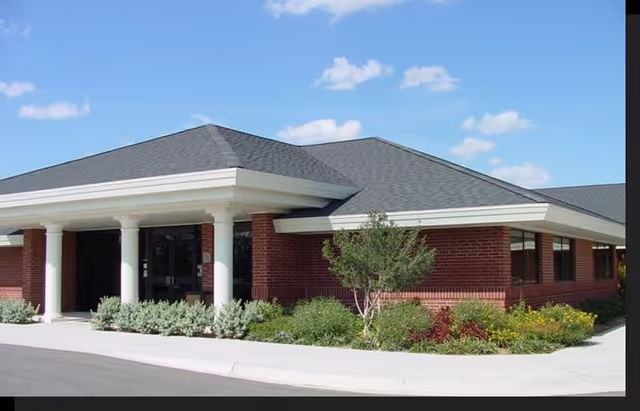 Exterior view of a single-story brick building with a gray shingled roof, white columns at the entrance, and surrounding landscaping with bushes and small trees under a blue sky with a few clouds.