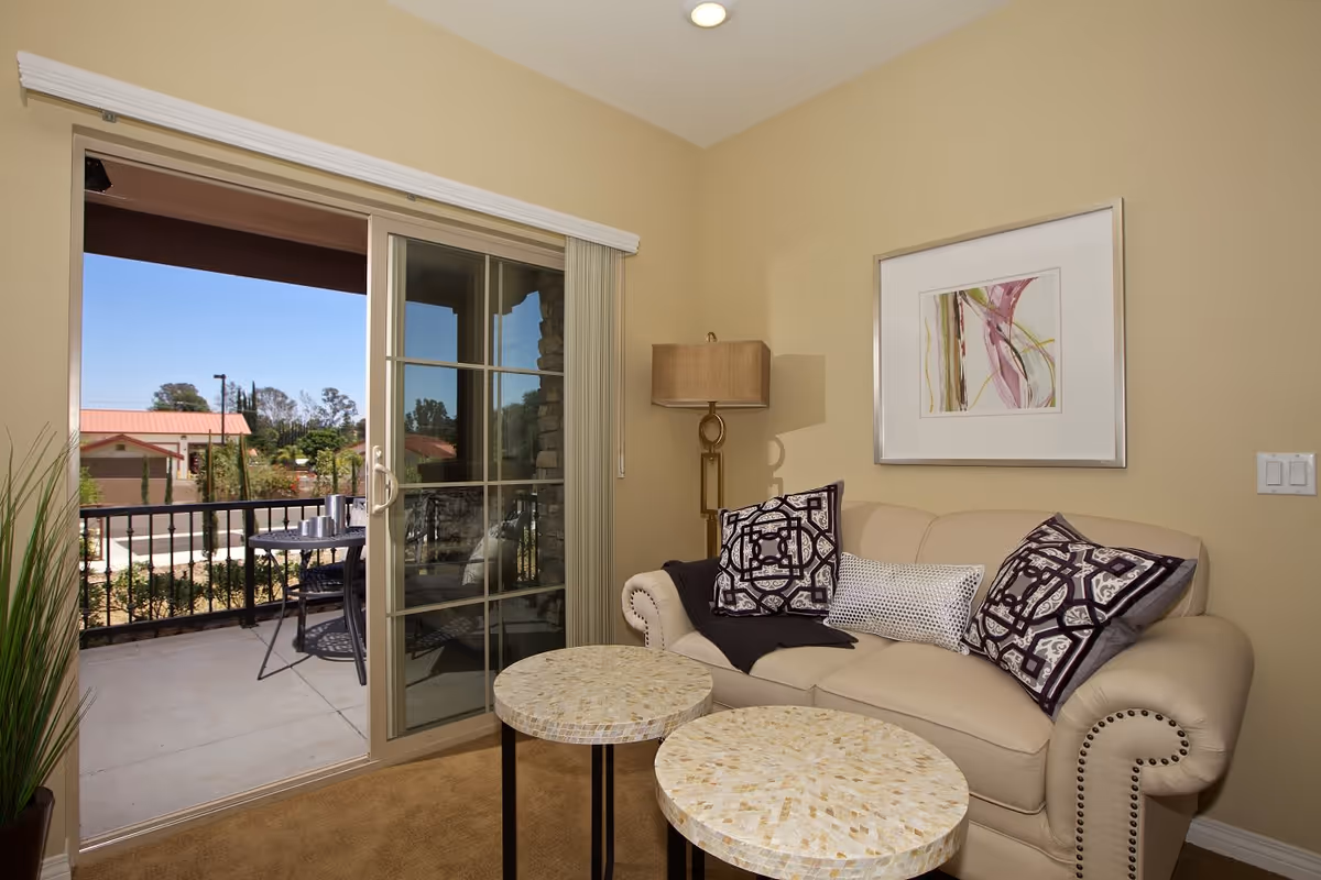 A cozy living room area with a beige couch adorned with three decorative pillows and a dark throw blanket. In front of the couch are two round mosaic-tiled coffee tables. To the right of the couch is a tall floor lamp with a beige shade. A framed abstract artwork hangs on the wall above the couch. Sliding glass doors open to a balcony with a small round table and two chairs, overlooking a residential area with trees and buildings under a clear blue sky.