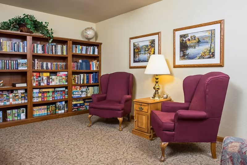 Cozy seating area with two purple wingback chairs, a wooden side table with lamp, and a bookshelf filled with books and games.