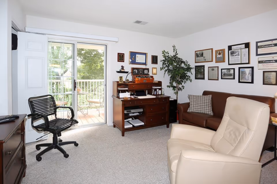 Bright living room/office area with a sliding glass door to a balcony, chairs, a wooden desk, and framed awards on the wall.