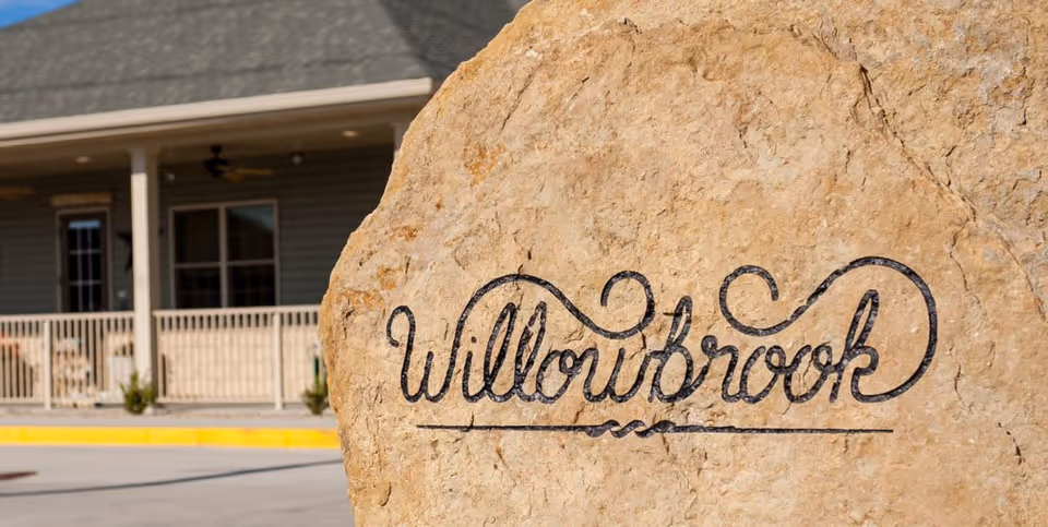 A large rock with the word 'Willowbrook' artistically engraved on it, positioned in front of a building with a porch and railing.