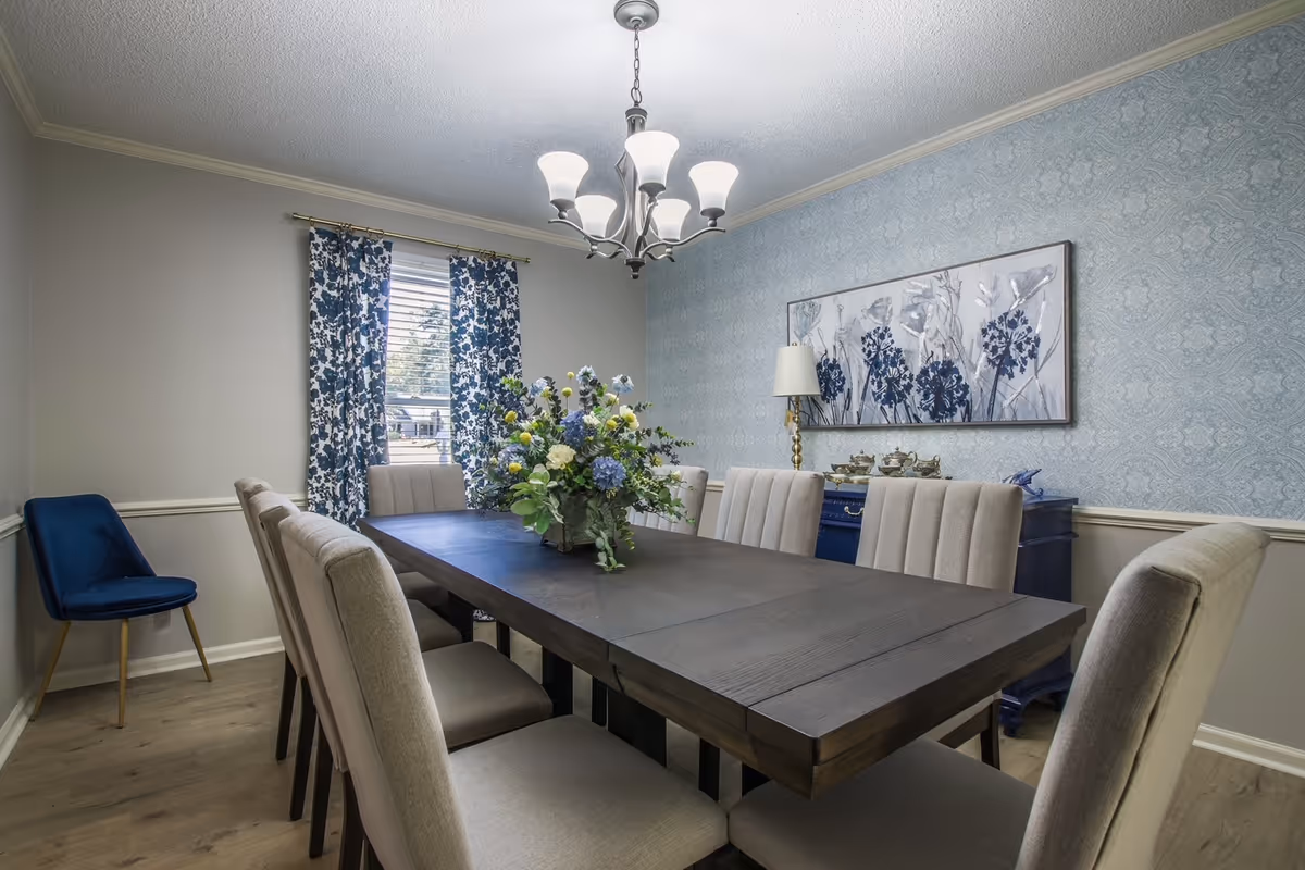 A dining room with a long dark wooden table surrounded by eight beige upholstered chairs. A large floral centerpiece sits on the table. The room has light blue patterned wallpaper on one wall, a window with blue and white floral curtains, a blue sideboard with a lamp and decorative items, and a modern chandelier hanging from the ceiling.