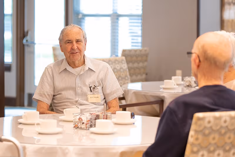 Two elderly men sitting at a round dining table in a well-lit room with cups and saucers on the table. One man is facing the camera and smiling, while the other is seen from behind. The room has patterned chairs and large windows with blinds.