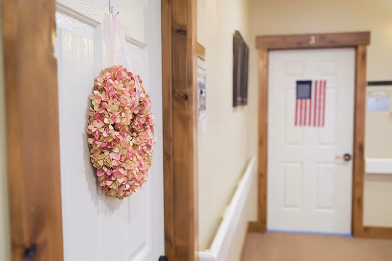 Hallway inside a senior living facility with a door in the foreground decorated with a pink floral wreath and another door with an American flag at the end.
