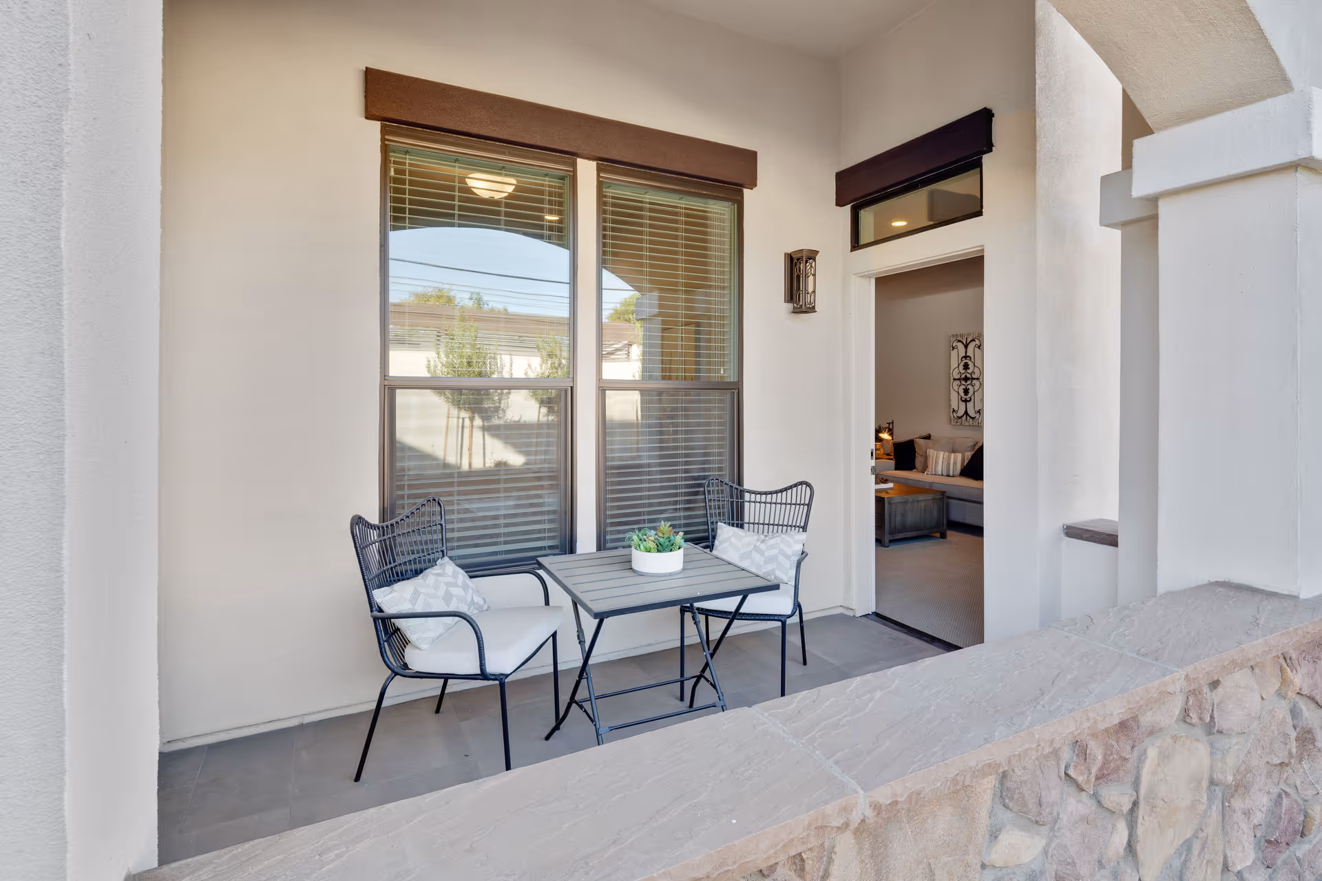 A small outdoor patio area with two black metal chairs with white cushions and a small square table with a potted plant on top. The patio is adjacent to a building with beige walls and large windows with blinds. Through an open doorway, a living room with a sofa and decorative wall art is visible.