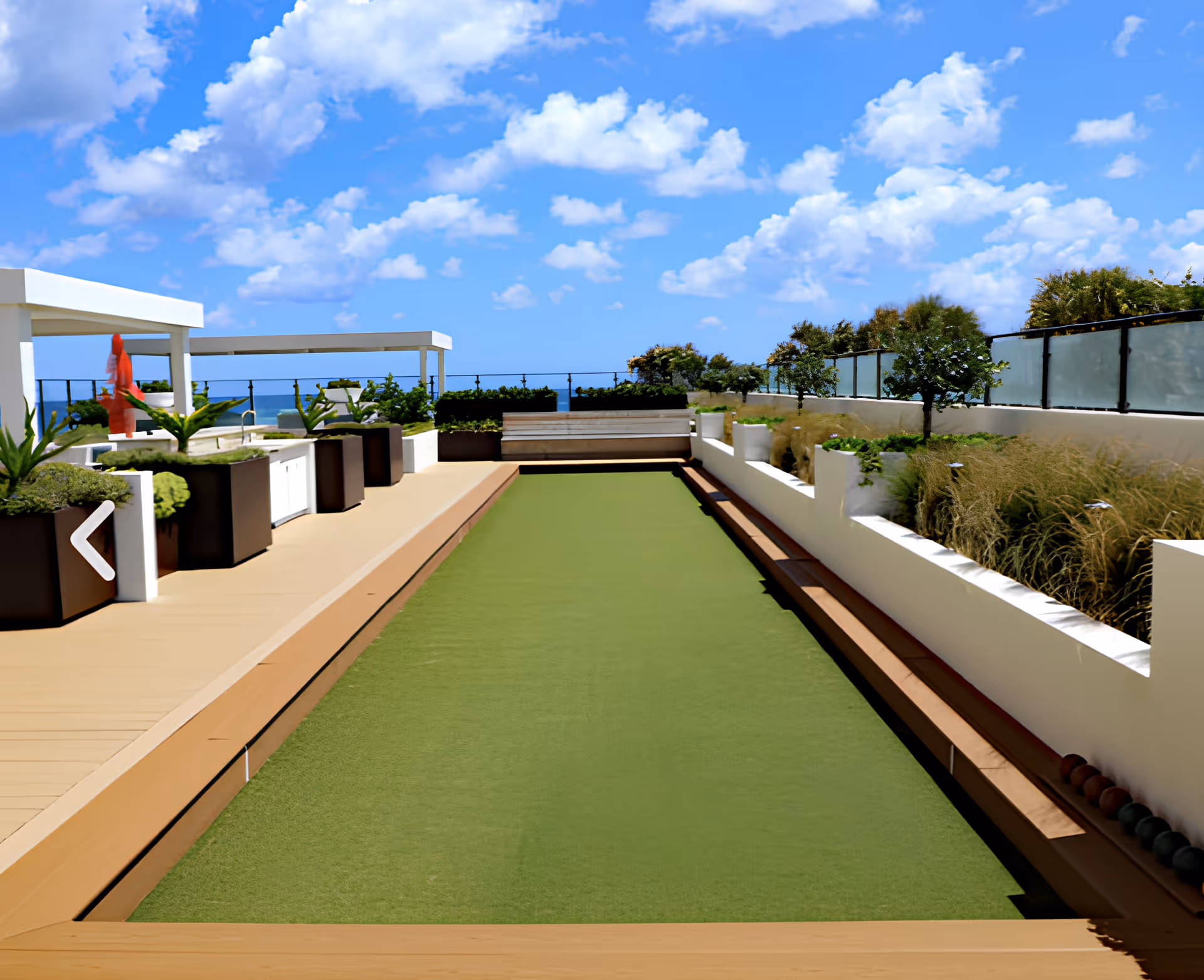 Outdoor bocce ball court with green playing surface, surrounded by wooden benches and planters with greenery. The area is on a rooftop with glass railing and a view of the blue sky with scattered clouds.