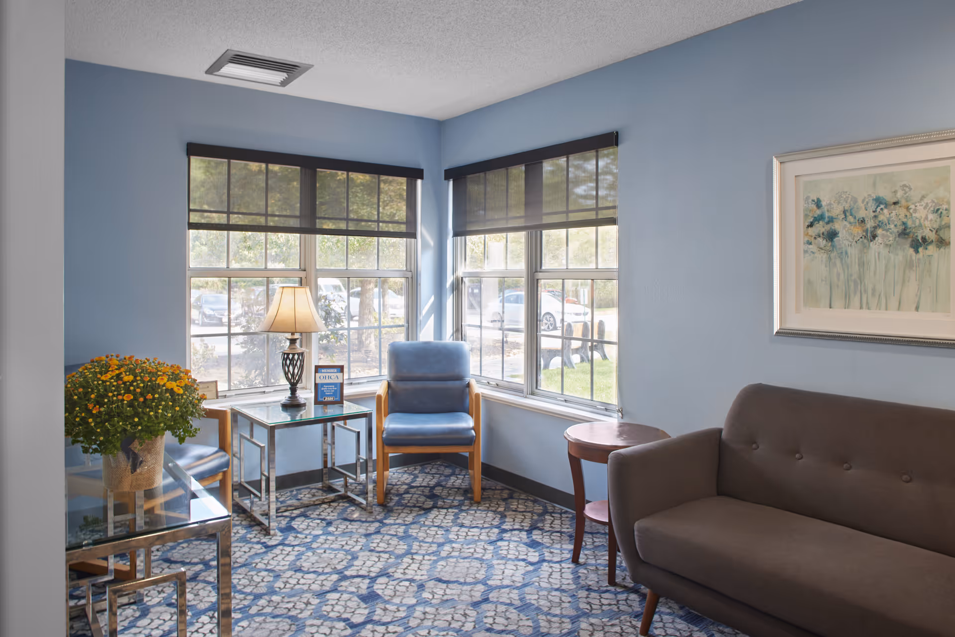 A cozy sitting area with light blue walls and large windows letting in natural light. The room features a blue armchair, a brown sofa, a side table with a lamp, a small round wooden table, a glass table with a potted plant, and a framed floral painting on the wall. The floor is covered with a patterned blue and beige carpet.