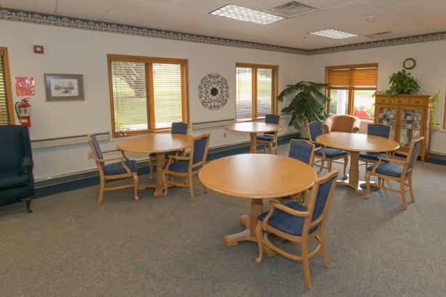 Communal dining room with four round wooden tables and blue-upholstered chairs, windows, plants, and a display cabinet.