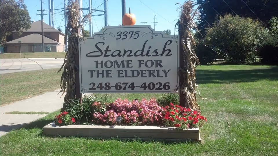 Outdoor sign for Standish Home For The Elderly with the address number 3375 and phone number 248-674-4026, surrounded by flowers and two dried cornstalks on either side, with a pumpkin on top of the sign.