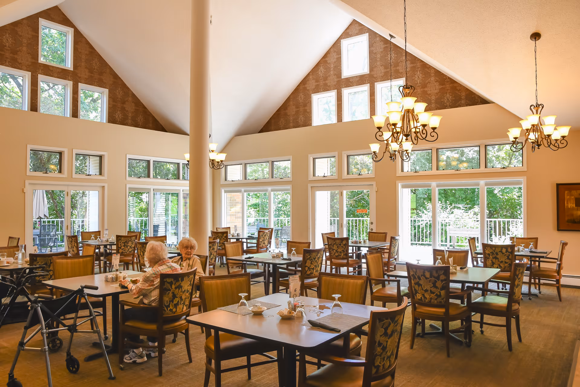 Bright senior living dining room with multiple tables and chandeliers, large windows, and two elderly women seated at a table.
