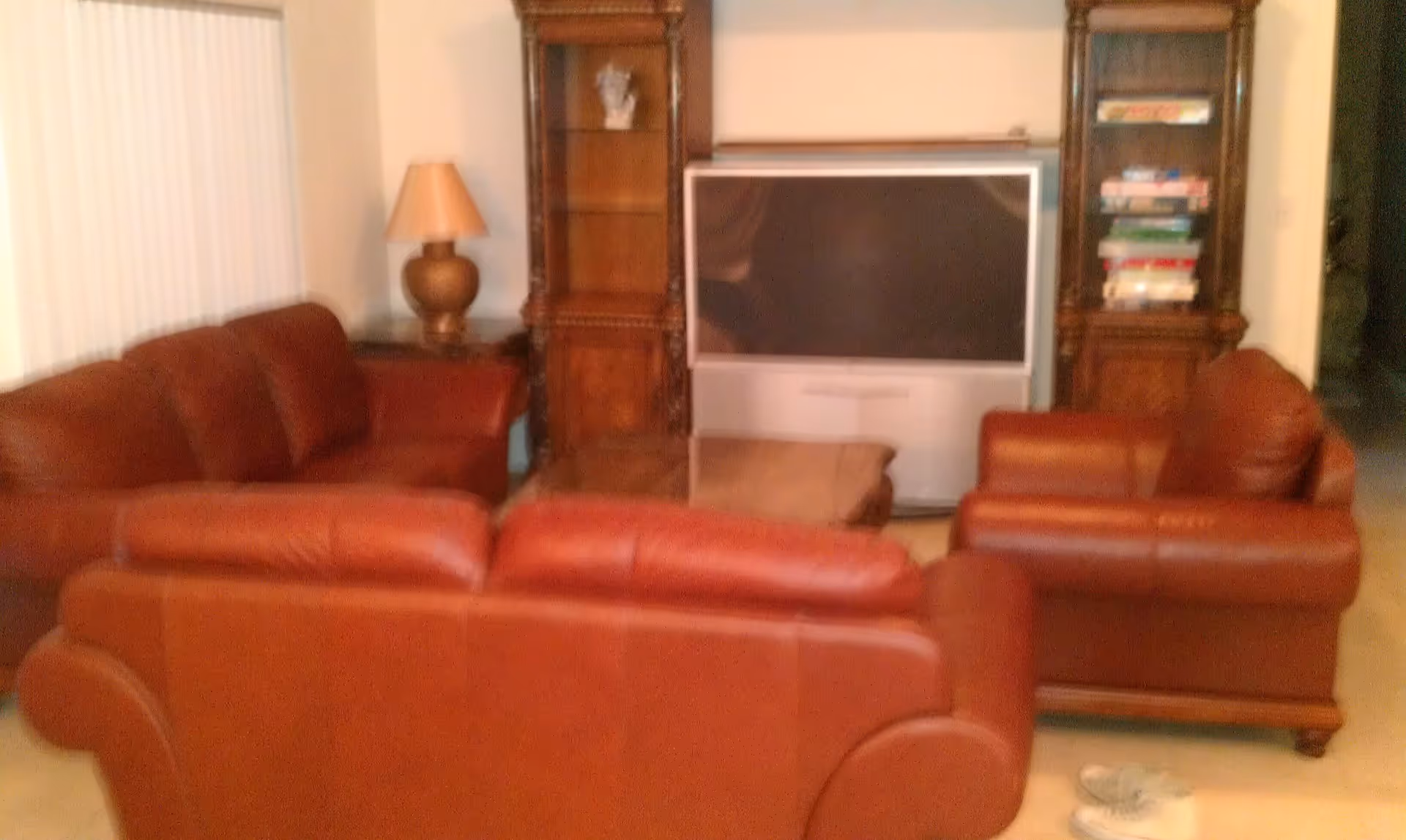 A living room with three red leather sofas arranged around a wooden coffee table. There is a large television set against the wall, flanked by two wooden shelving units, one with books and the other with a decorative item. A table lamp is on a side table next to one of the sofas. A pair of white shoes is on the floor near the sofas.